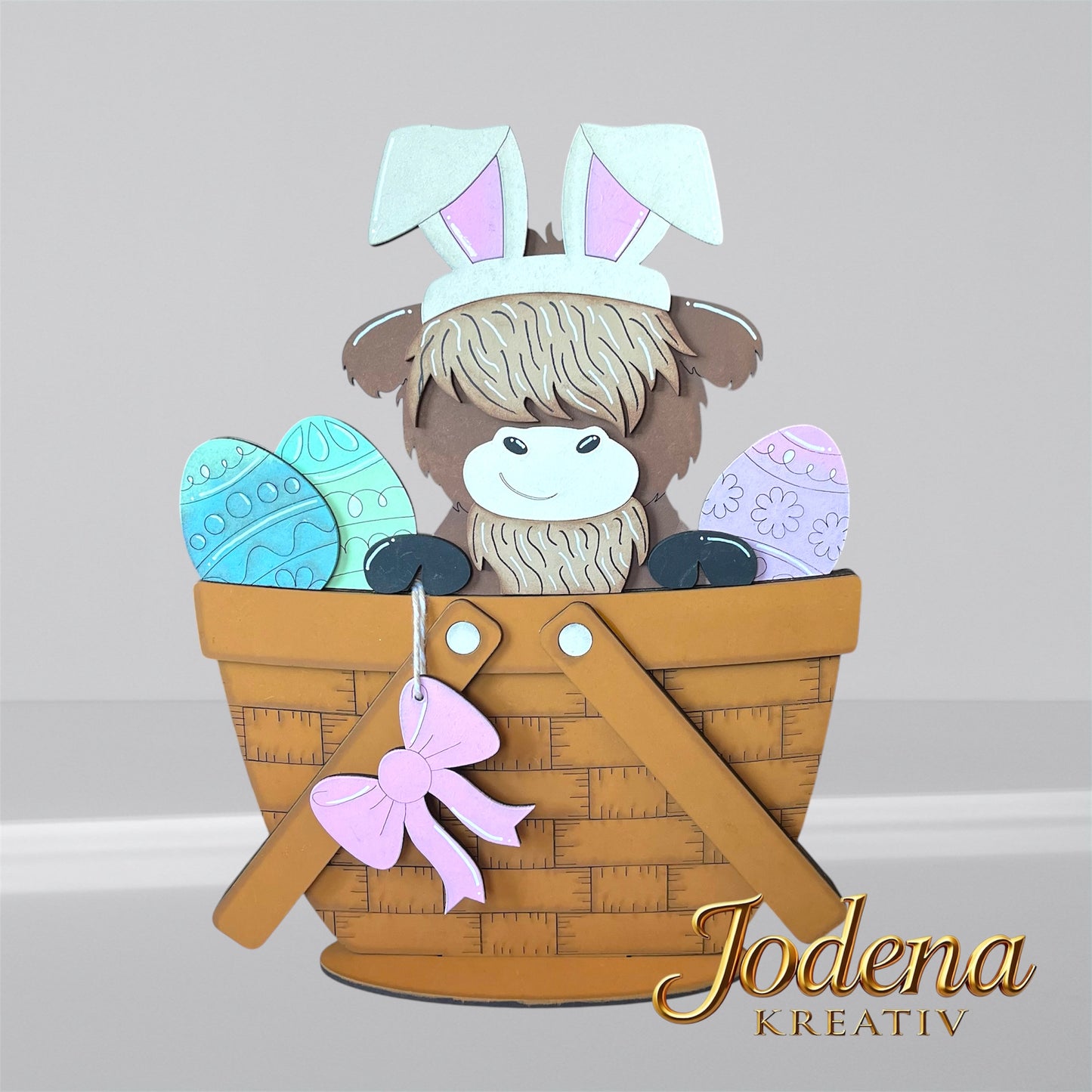 Austauschbarer Korbeinsatz mit Hochlandkuh und Hasenohren, sitzend neben bunten Ostereiern im braunen Dekokorb
