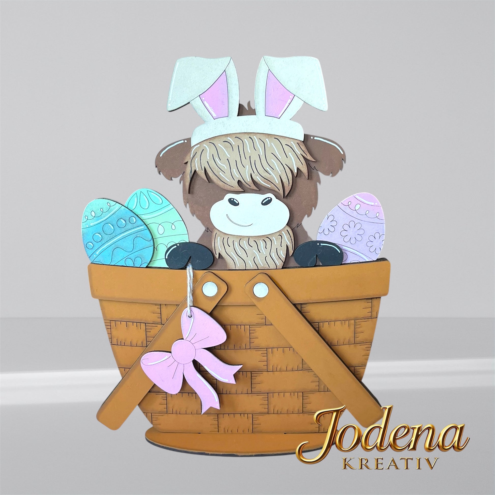 Austauschbarer Korbeinsatz mit Hochlandkuh und Hasenohren, sitzend neben bunten Ostereiern im braunen Dekokorb