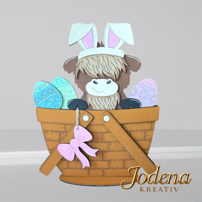 Austauschbarer Korbeinsatz mit Hochlandkuh und Hasenohren, sitzend neben bunten Ostereiern im braunen Dekokorb
