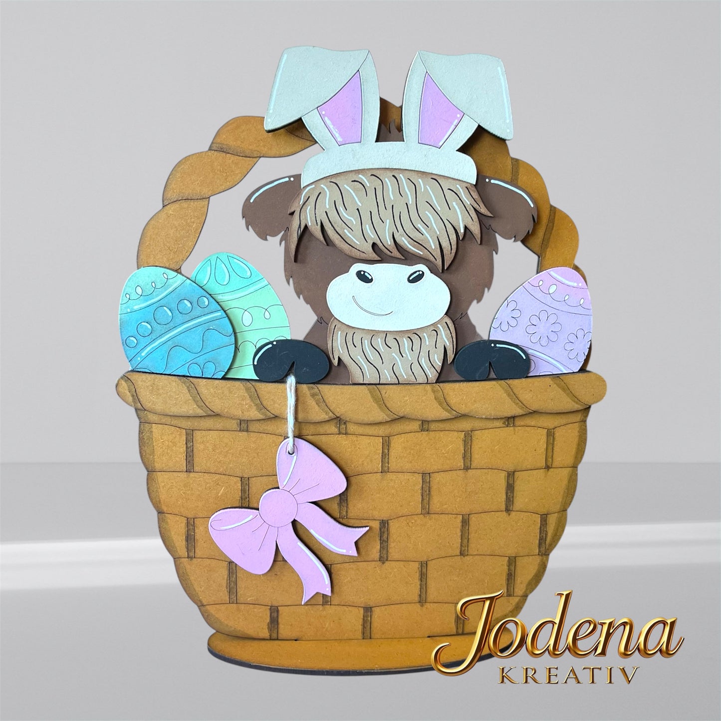 Austauschbarer Korbeinsatz mit Hochlandkuh und Hasenohren, sitzend neben bunten Ostereiern im braunen Osterkorb