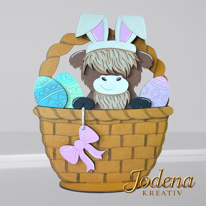 Austauschbarer Korbeinsatz mit Hochlandkuh und Hasenohren, sitzend neben bunten Ostereiern im braunen Osterkorb