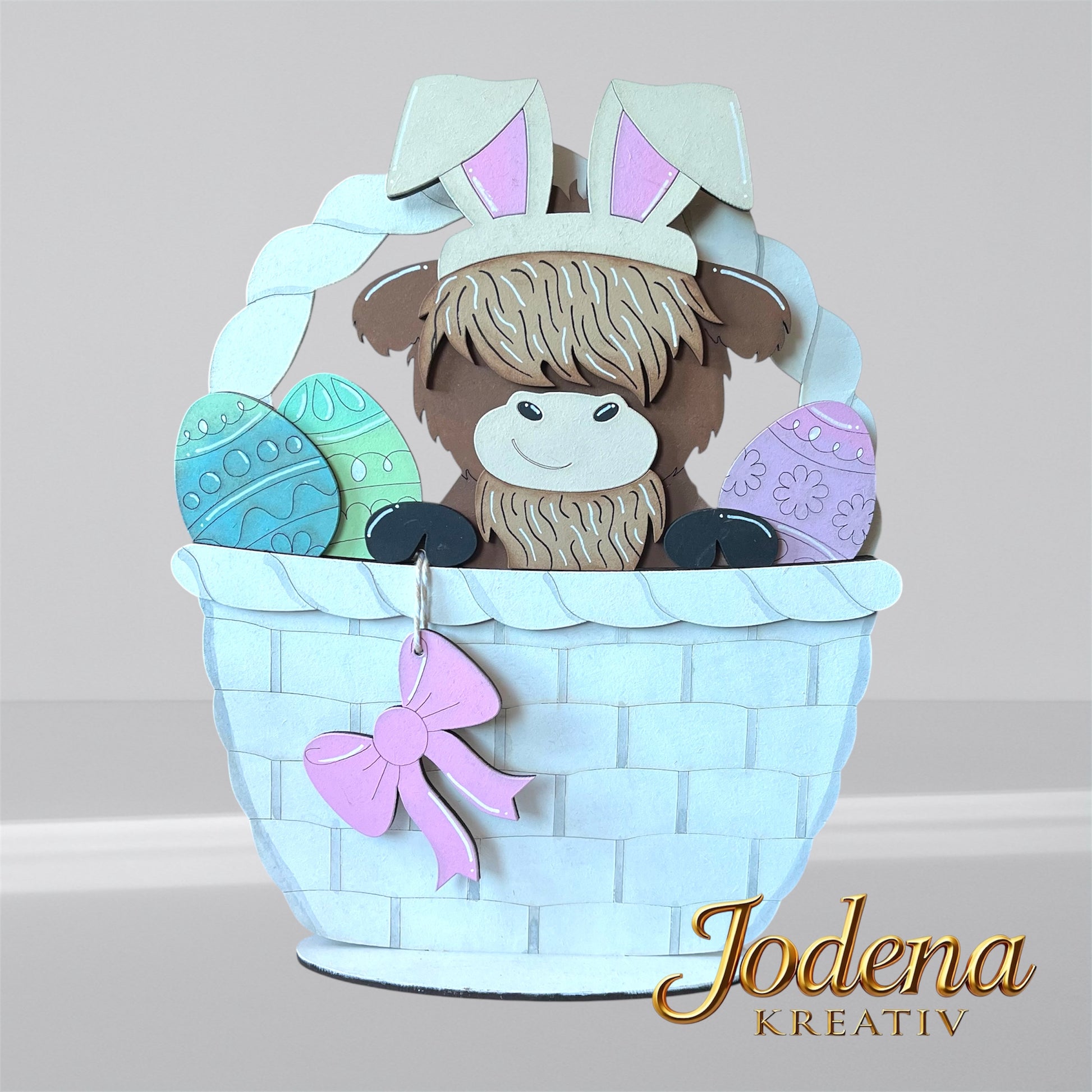 Austauschbarer Korbeinsatz mit Hochlandkuh und Hasenohren, sitzend neben bunten Ostereiern im weißen Osterkorb