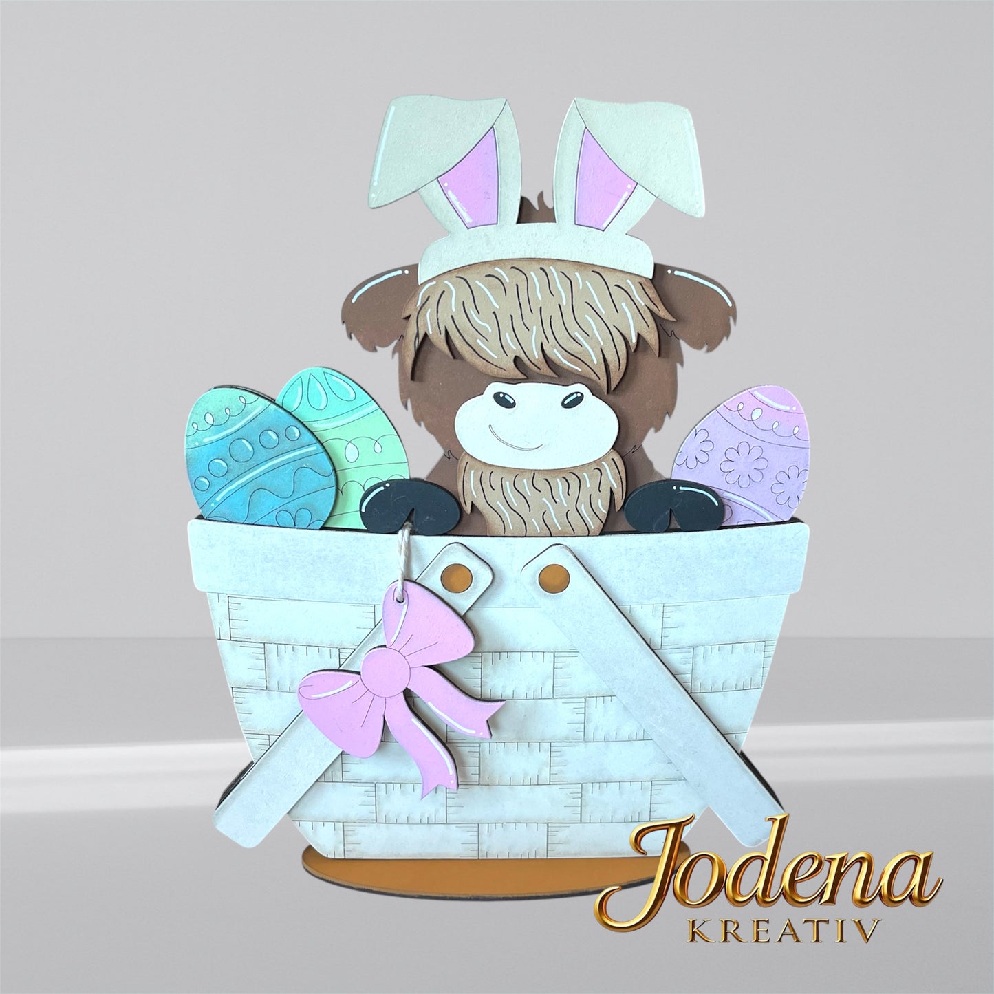 Austauschbarer Korbeinsatz mit Hochlandkuh und Hasenohren, sitzend neben bunten Ostereiern im weißen Dekokorb