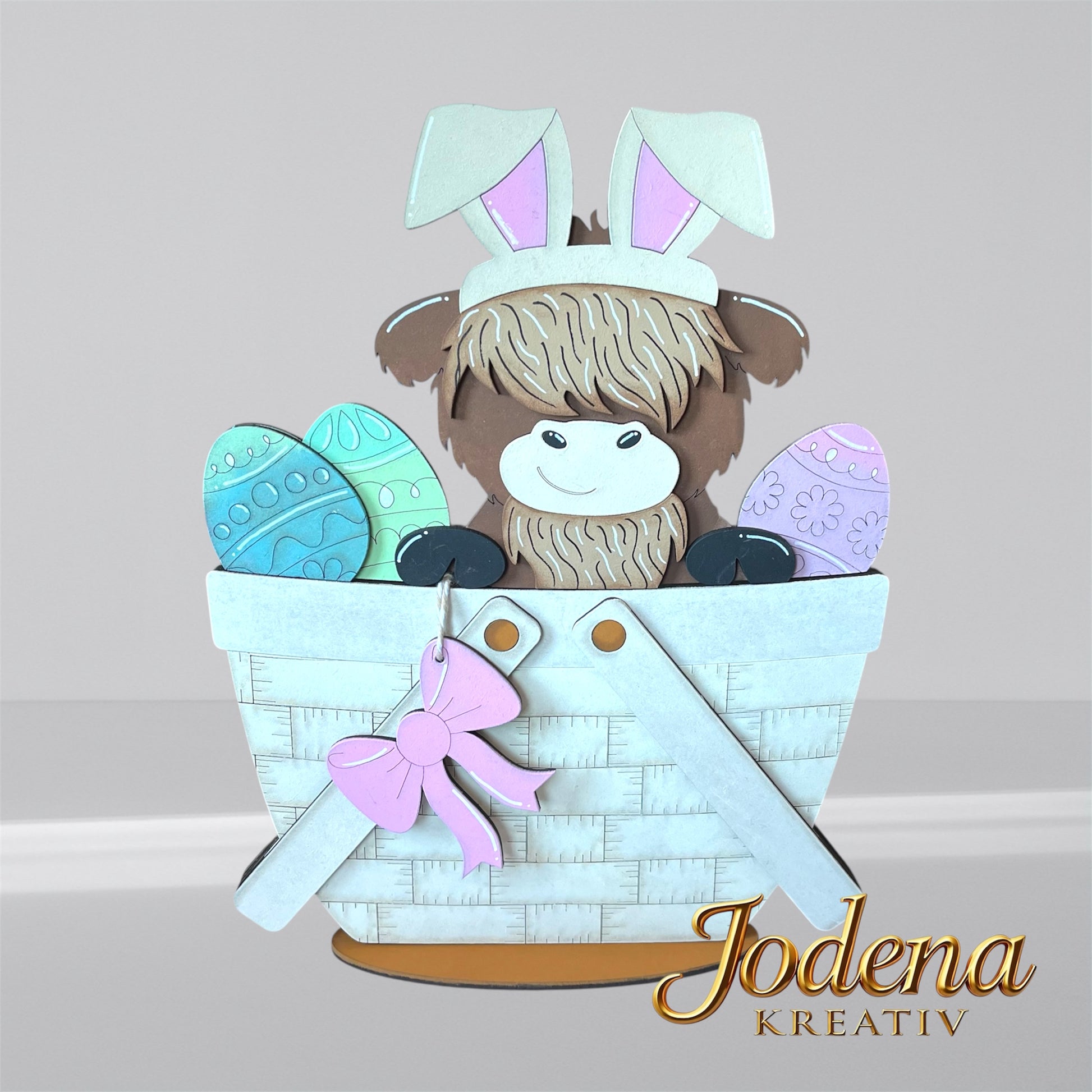 Austauschbarer Korbeinsatz mit Hochlandkuh und Hasenohren, sitzend neben bunten Ostereiern im weißen Dekokorb